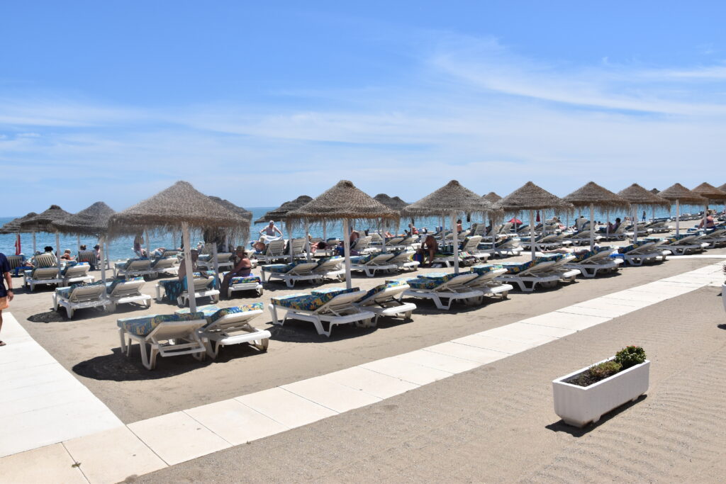 Rows of beach sunbeds and straw umbrellas on the sand next to the ocean and the sea promenade in Fuengirola.