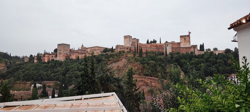 View of the historic Alhambra palace walls and towers in Granada viewed from the city below against a dramatic sky.