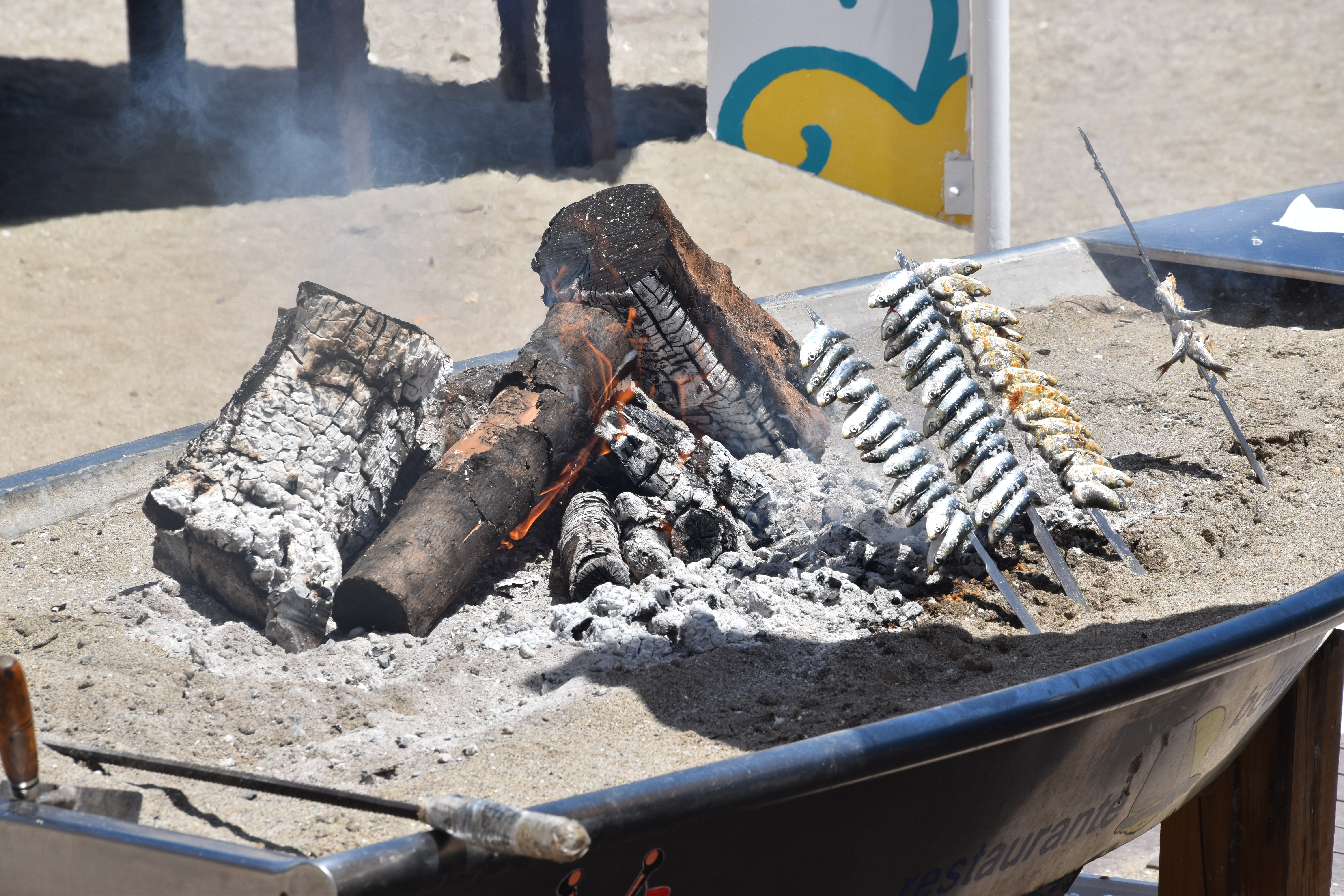 Rows of fresh sardines on skewers (espetos) grilling over an open olive-wood fire inside a traditional, converted fishing boat on a sandy Costa del Sol beach in Spain.