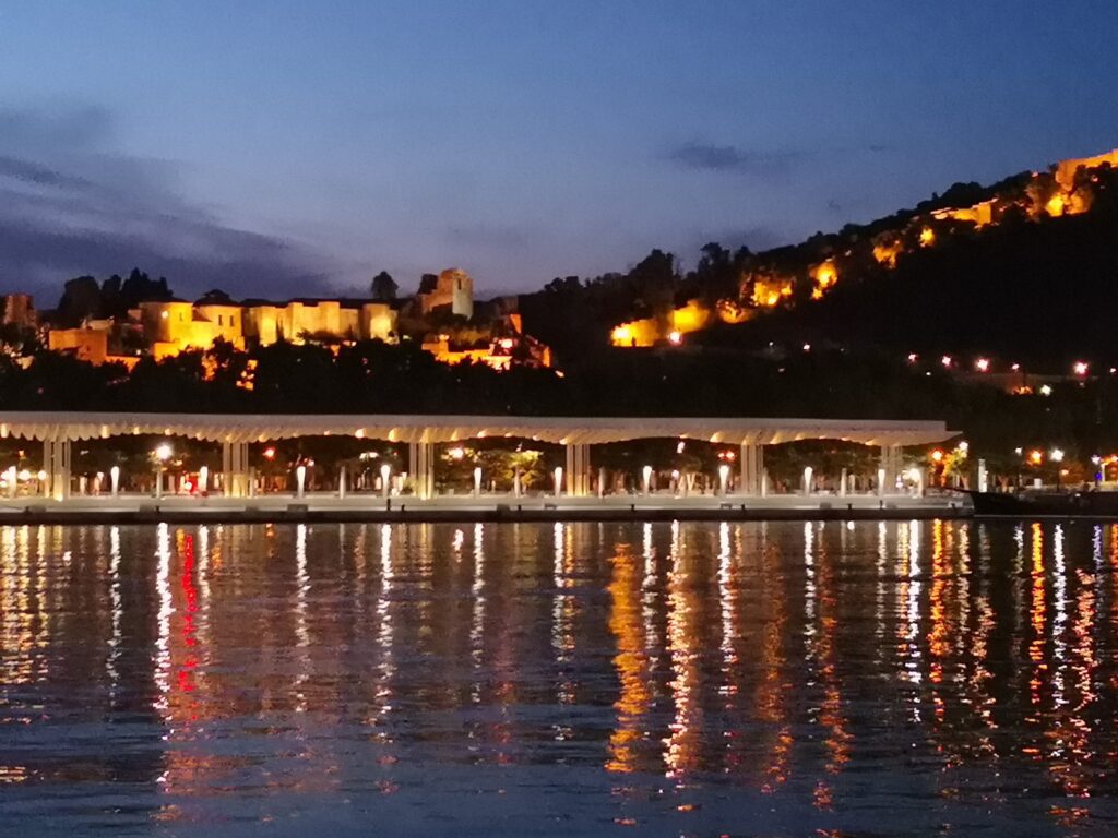 Oplyst Alcazaba-fæstning og Málaga havn om natten med lysrefleksioner i vandet ved Muelle Uno